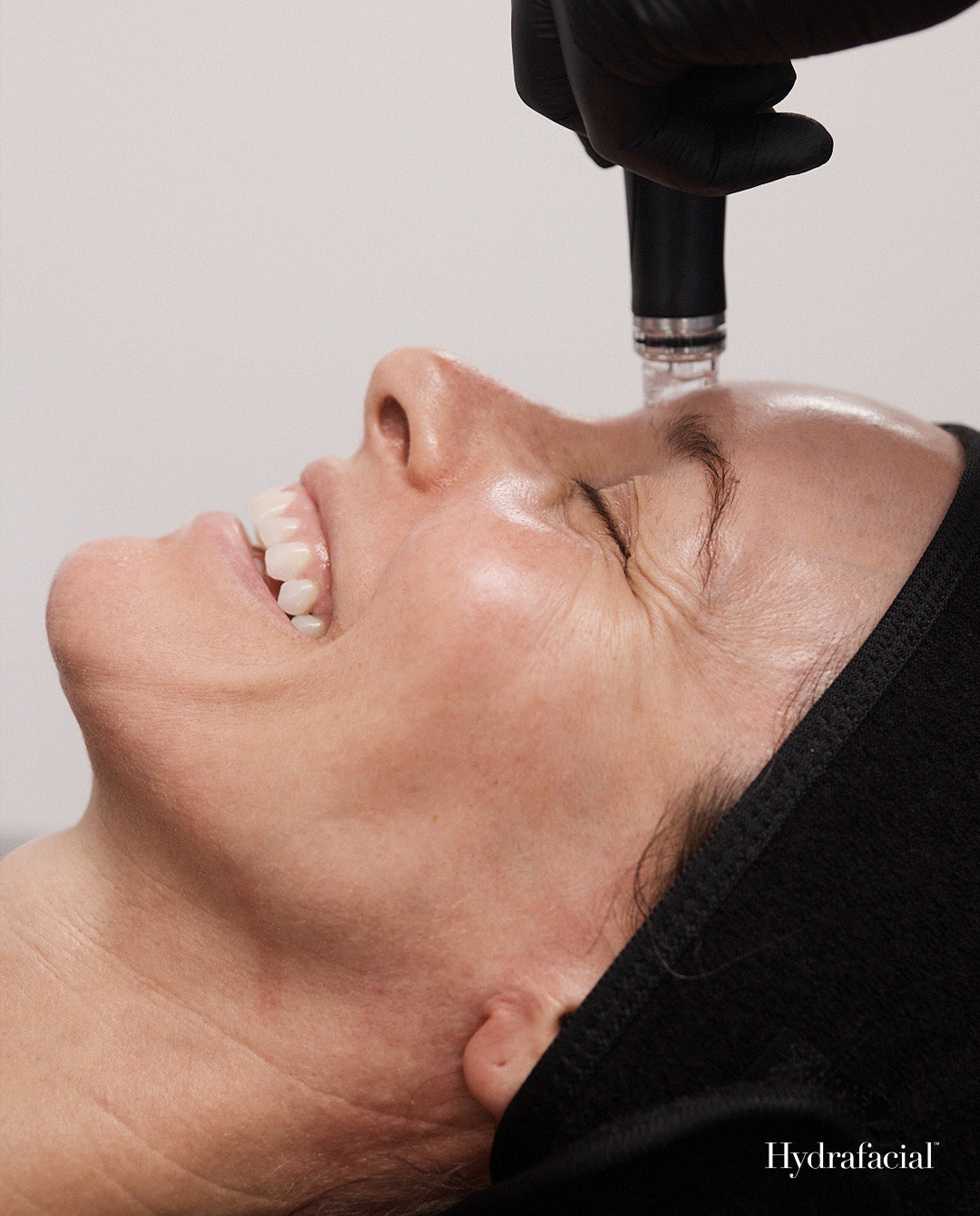 A close-up of a woman lying down with her eyes closed while a gloved hand applies a clear facial treatment to her forehead. She is wearing a black headband, and the word 'Hydrafacial' appears in the bottom right corner.